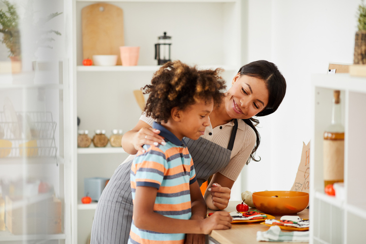Positive young black mother touching shoulder of son while talking to him in kitchen, they preparing treats for Halloween party